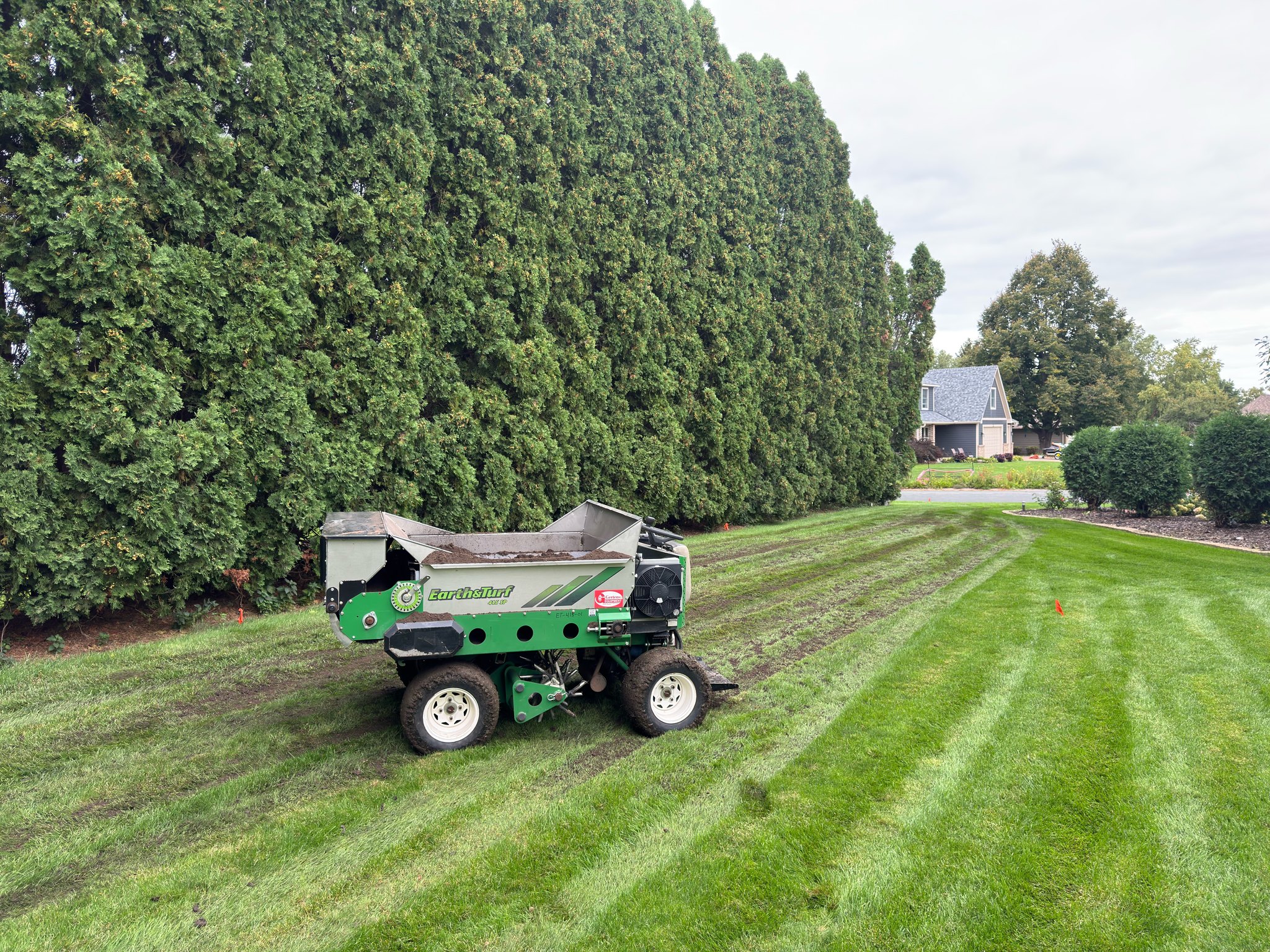 Topdressing machine applying material across a residential lawn beside tall evergreen trees.